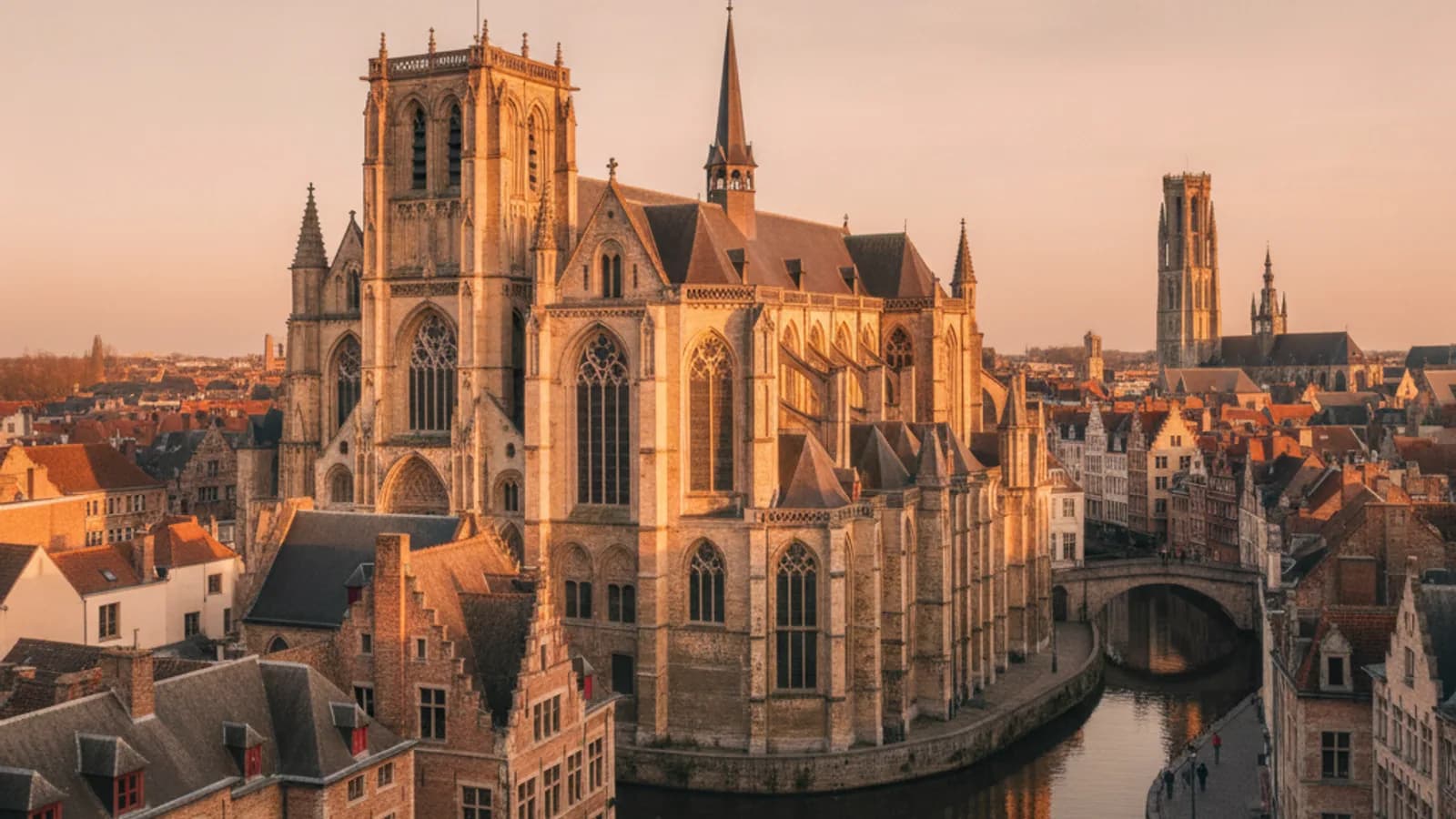 Vue panoramique sur les toits de Gand au coucher du soleil, avec le Beffroi, la Cathédrale Saint-Bavon et l'Église Saint-Nicolas se détachant sur l'horizon, et les quais de la Lys en contrebas.